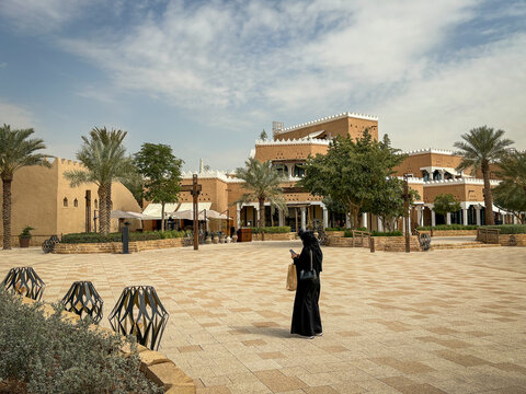 A woman in a black abaya crosses a paved square before a restored mud brick complex, Riyadh, Riyadh, Saudi Arabia
