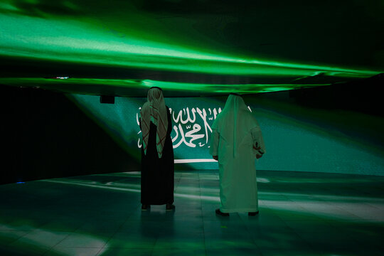 Two visitors in traditional dress stand beneath a screen projecting the Saudi flag, Riyadh, Riyadh, Saudi Arabia