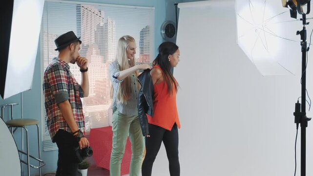 A male photographer directs a group of four female models changing clothes and posing inside a professional photo studio with city skyscrapers visible through a window.