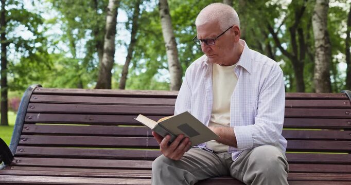 Senior man reading book on park bench. Nature setting with trees supports relaxation and learning outdoors. Casual clothing and glasses suggest comfort, wisdom. Calm senior reading idea now.