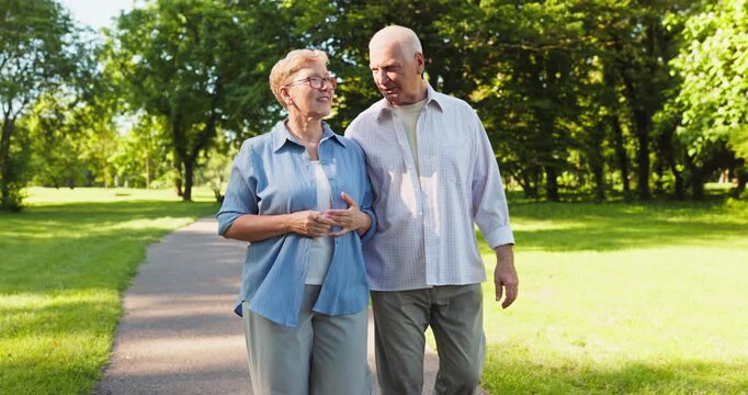Senior couple walking outdoors in a park. They walk arm in arm on a sunny path amid green trees, signaling wellness, relationship and positive aging in retirement. Active life and love win.