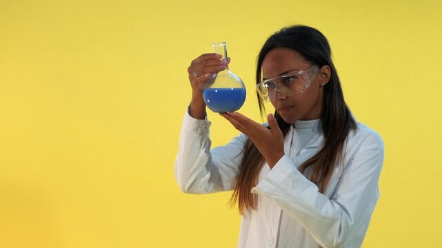 Black woman wearing lab coat and safety glasses carefully holding a glass flask with blue liquid against a yellow background in a studio setting.