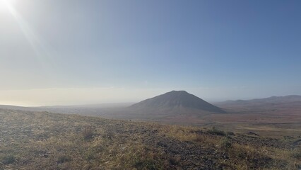 Misty volcanic landscape with a lone cone-shaped volcano rising above a dry, barren plain under a hazy blue sky, a remote desert wilderness in soft morning light © Konic