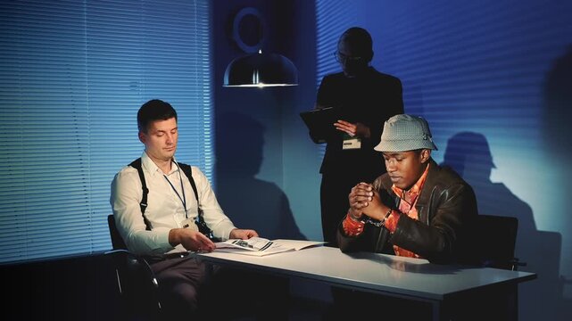 Three men engaged in a serious discussion around a table covered with documents in an interrogation room with blinds and dramatic lighting.