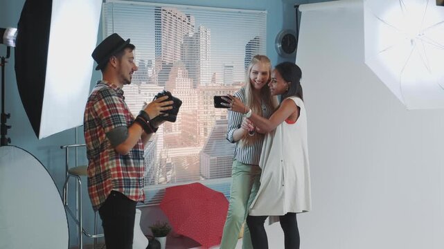 Three young women pose with smartphones while a man photographs them in a professional indoor photo studio with urban cityscape backdrop.