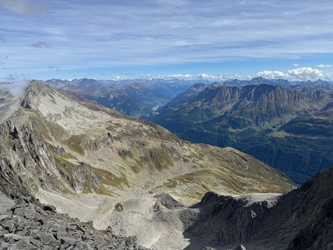 High-altitude panoramic mountain view in the Swiss Alps near Realp, Uri, showing jagged rocky peaks, dramatic ridges, alpine valleys and cloud layers under a deep blue sky in the wild landscape