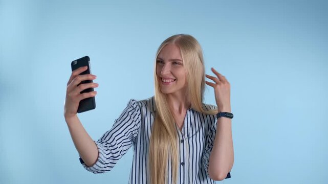A young blonde woman wearing a striped shirt takes selfies with a black smartphone against a plain light blue indoor background. Various expressions shown.