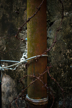 Close up of a rusty metal pipe wrapped with barbed wire and cables on a dark weathered wall, representing security, aging infrastructure, corrosion, deterioration, maintenance, and urban decay.