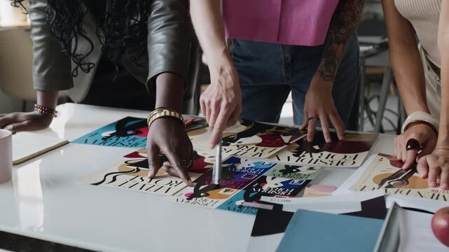 Diverse team of young adult female designers reviewing layout prints and posters on table during creative meeting. Clip useful for advertising agency workflow and graphic design marketing