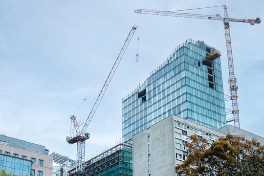Workers receive hoisted glass unit at open floor of glazed tower flanked by cranes. Urban densification, new tower next to old block, changing city skyline