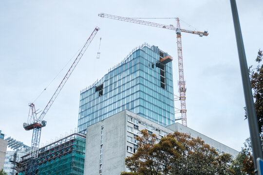 Tower cranes lift panel onto unfinished glass office high rise beside older concrete apartment block. New office tower construction, office vacancy rates, commercial real estate market