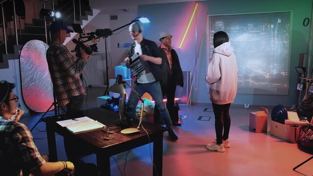 A male actor aims a prop gun at a female actress inside a large industrial film studio while a crew member observes nearby amid professional production equipment.