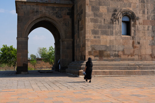 A woman dressed in black walks across a paved courtyard beside an ancient stone church with a large arched entrance and ornate medieval window in Armenia. 