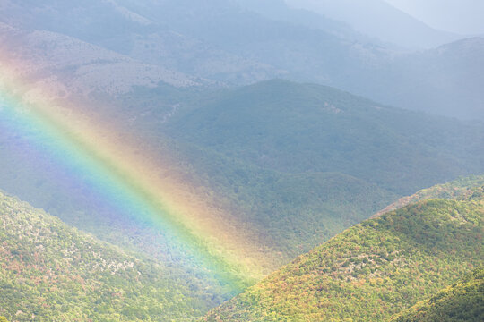 A vibrant rainbow arcs across a misty valley covered in lush green mountains. Beautiful atmospheric phenomena inspire deep feelings of hope, natural harmony and peaceful spiritual renewal.