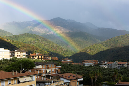 A bright rainbow arcs over green mountains and a rural village with brick roofs after a storm. Such dramatic weather phenomena inspire deep feelings of hope, peaceful renewal and community harmony.
