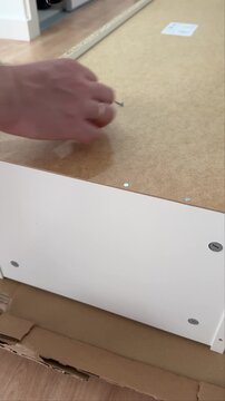 Hand hammering a nail into the fiberboard back of a white cabinet, vertical close-up. Vertical close-up of a man driving a nail into a cabinet back panel with a hammer.