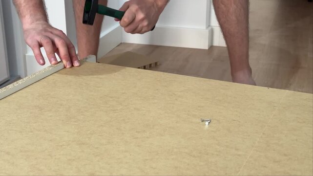 Man confidently driving two nails into the fiberboard back of a cabinet lying on the floor. Man hammers two nails into a cabinet back panel lying on the floor at home.