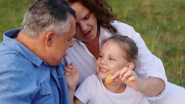 Close up cheerful little girl feeding dad biscuit outdoors. Loving mother watching with smile. Happy family sharing snack, having fun together during cozy summer picnic in park. Leisure time in nature