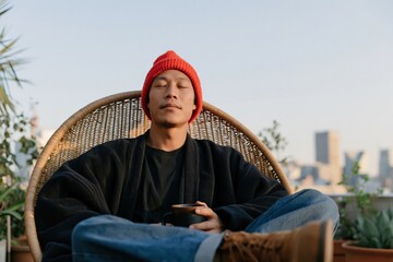 Obraz premium Rooftop portrait of relaxed man in red beanie holding a coffee mug for lifestyle banner and calm urban background design