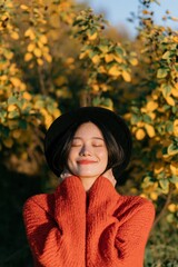 Obraz premium Young woman in a cozy orange sweater and black hat enjoying autumn sunlight among yellow leaves for seasonal portrait background and lifestyle design