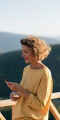Obraz premium Middle aged woman with curly hair in a yellow sweater checking phone on a mountain balcony holding a glass — lifestyle outdoor portrait background for banner and wallpaper