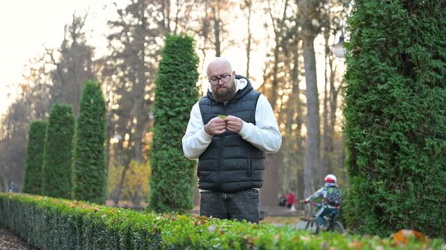 Man stands among greenery in peaceful park, skillfully playing harmonica while sunlight filters through trees. Autumn leaves gently fall around him, creating a tranquil atmosphere.