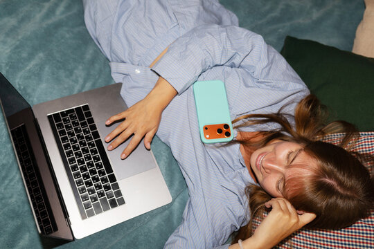 Woman in pajamas in bed with a laptop and a phone in a cozy room