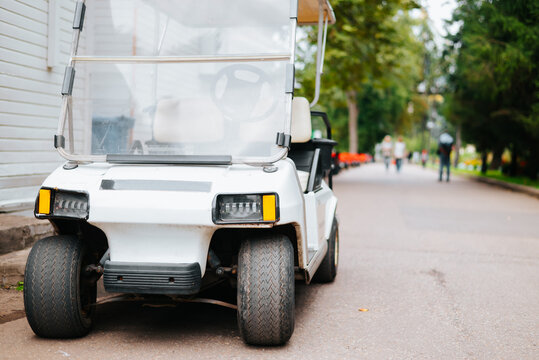 A white electric golf cart is parked on a pathway surrounded by trees and greenery. People can be seen in the background walking or engaging in activities in a recreational setting