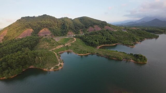 Aerial drone footage moves along a mountain reservoir past a wooded shoreline and dense green vegetation beneath warm evening light.