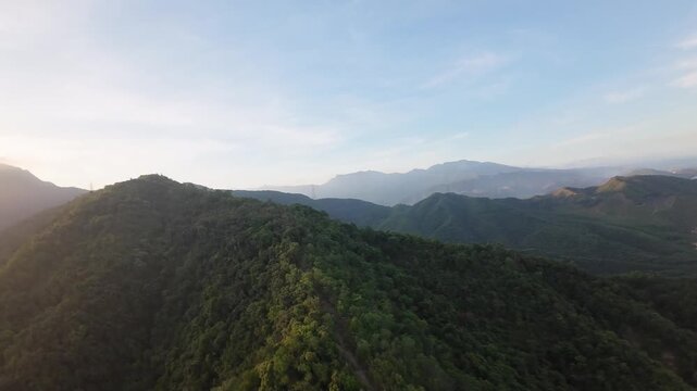Aerial drone footage glides above dense forested mountains and layered ridges illuminated by warm evening sunlight.