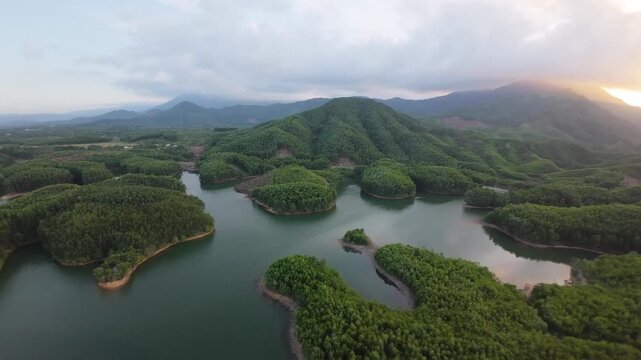 Aerial drone footage moves along a mountain reservoir past a wooded shoreline and dense green vegetation beneath warm evening light.