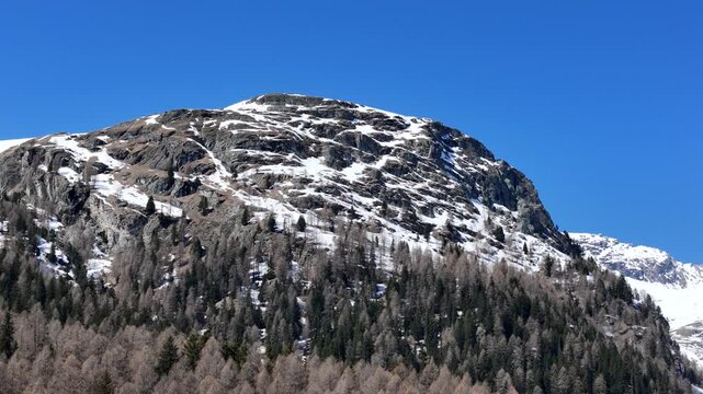 patchy snow streaks rugged rock face broad summit towers above forested larch pine slopes clear blue sky aerial drone reveal julier pass mountain pass graubunden switzerland swiss alps high altitude