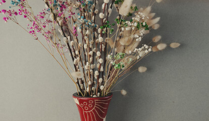 A vibrant arrangement of colorful dried flowers, featuring pussy willows and gypsophila, elegantly displayed in a decorative red vase with ethnic motifs against a neutral grey backdrop.  © Kos6ka