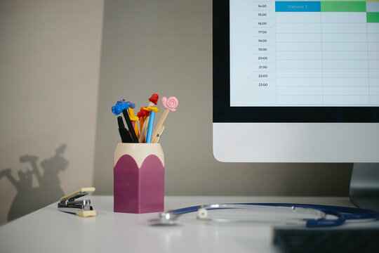 Pediatrician desk displaying stethoscope, computer monitor showing patient schedule, colorful pens. Image representing pediatric healthcare, suitable for clinic marketing