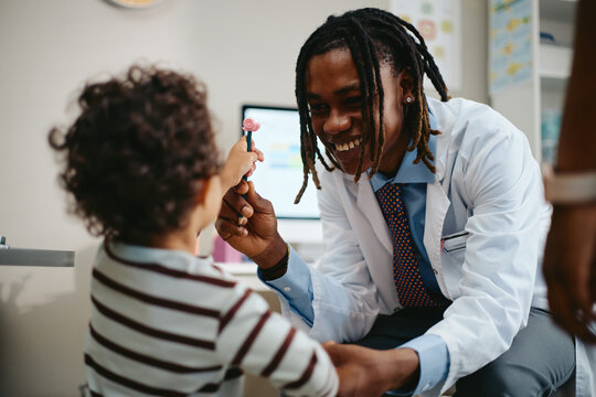Smiling African American young adult male pediatrician interacting with toddler patient during medical checkup in clinic. Image useful for pediatric healthcare and hospital marketing