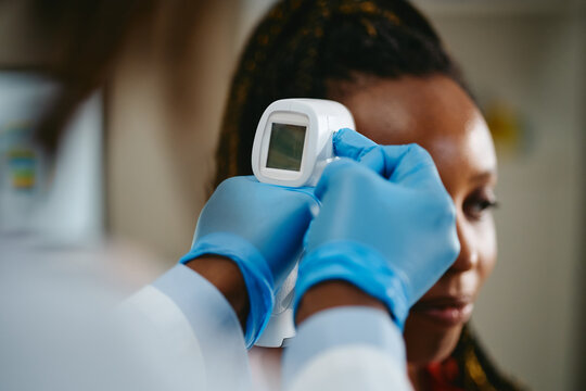 Doctor measuring temperature of young African American woman using infrared forehead thermometer in clinic. Image suitable for healthcare screening and fever checking concepts