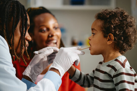 African American pediatrician examining toddler boy throat with wooden spatula beside mother in clinic. Medical checkup image suitable for healthcare website