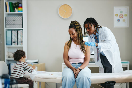 African American male doctor examining young adult Black woman with stethoscope while toddler child playing nearby. Image suitable for family healthcare and clinical medicine advertising