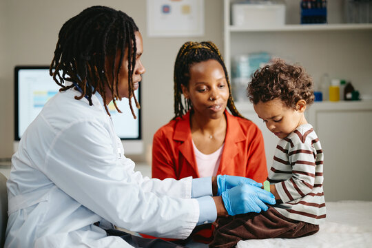 African American doctor applying bandage on arm of toddler boy near mother in clinic. Image ideal for pediatric healthcare, family medicine and medical clinic advertising