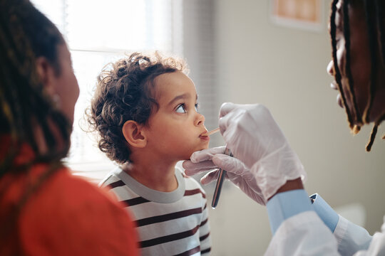 African American pediatrician examining toddler mouth with tongue depressor during clinic visit. Professional image suitable for pediatric healthcare and medical insurance campaigns