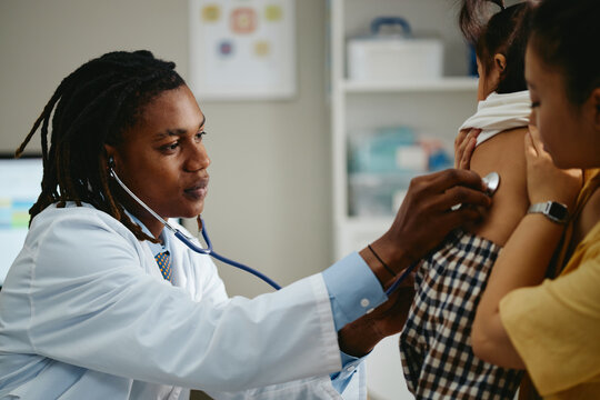 African American male pediatrician examining Asian child patient with stethoscope during clinical checkup. Pediatric healthcare concept for medical advertising and family clinic promotion