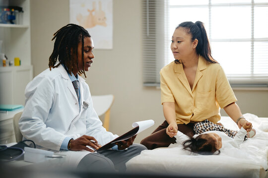 Young adult African American male pediatrician consulting Asian mother with baby child in clinic. Useful for pediatric healthcare campaigns and family insurance advertising