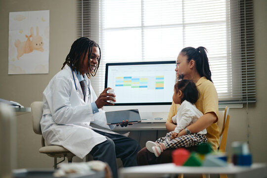 African American male doctor holding tablet while explaining medication to Asian mother holding toddler child. Healthcare image for pediatric clinics and medical consultations