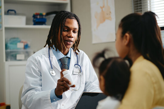 African American male pediatrician holding medicine bottle, explaining treatment to family in clinic. Concept representing healthcare marketing and child care editorial use