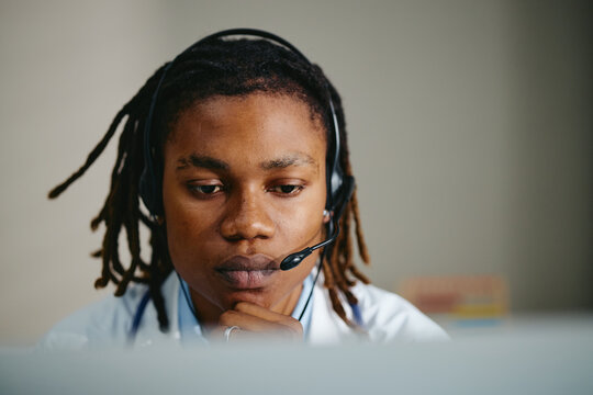 Young adult African American doctor wearing headset working on computer during online telehealth consultation. Image suitable for telemedicine, virtual healthcare, remote pediatric advice