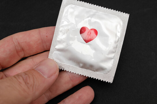 Blank condom in silver foil wrapper with red heart held in hand on black background