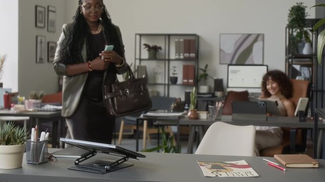 African American young adult businesswoman arriving at open office, greeting colleague, sitting at desk and opening laptop. Suitable for corporate communications and workplace video ads