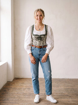 Smiling young    German woman wearing a traditional Alpine dirndl-inspired bodice and white embroidered blouse paired with modern blue jeans and sneakers.