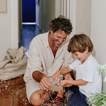 Christmas lights being wound onto a card by a father and son, glowing amber bulbs in a dark hallway for holiday and homeware campaigns