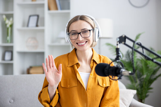 Podcaster and content creator, smiling woman with headphones and microphone waving hand for a live stream or online broadcast in a home office studio. Professional influencer recording audio content.